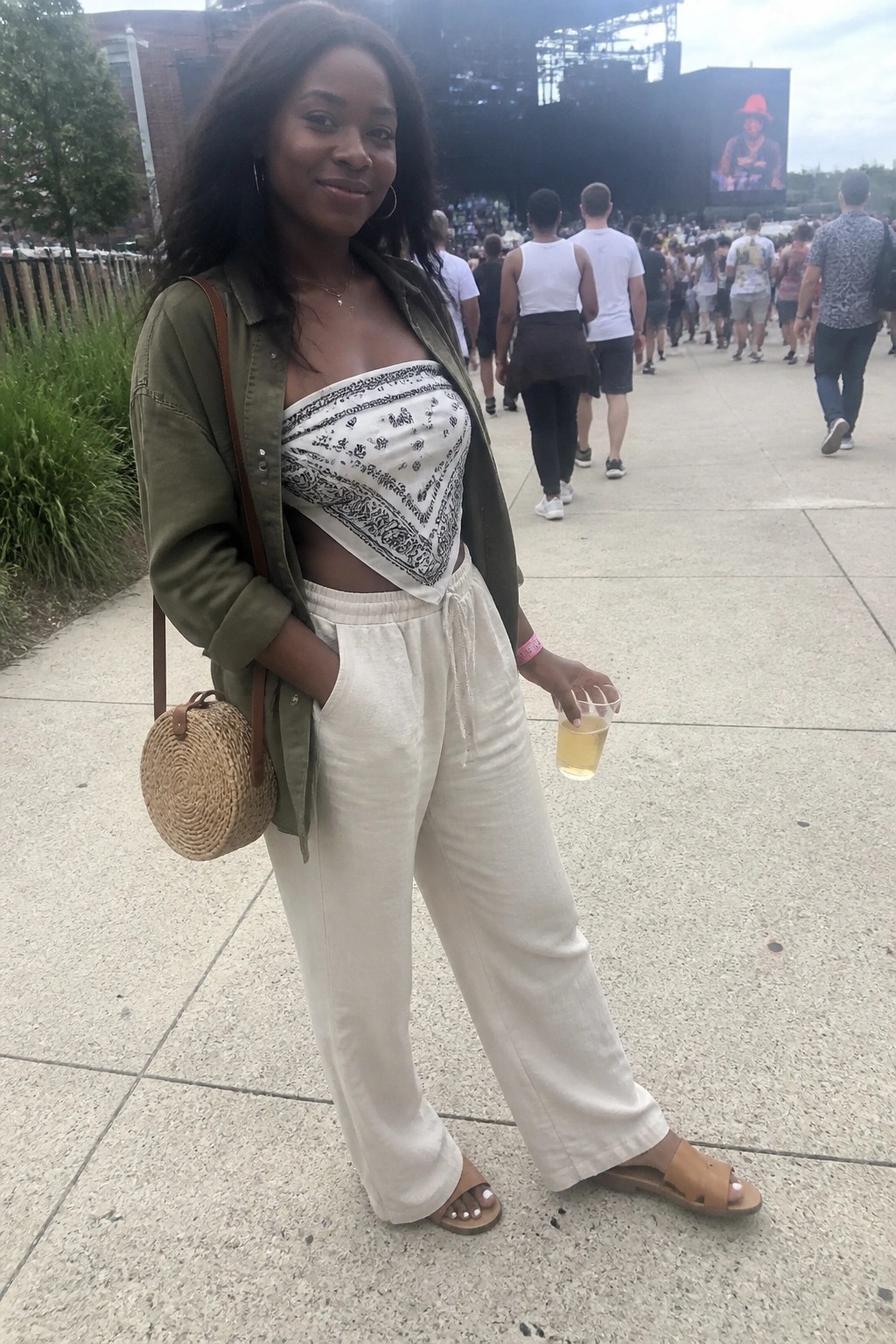 A woman wearing white wide-leg pants, an olive green button-up shirt open over a white bandana print bandeau top, holding a drink with a woven round bag at a concert venue.