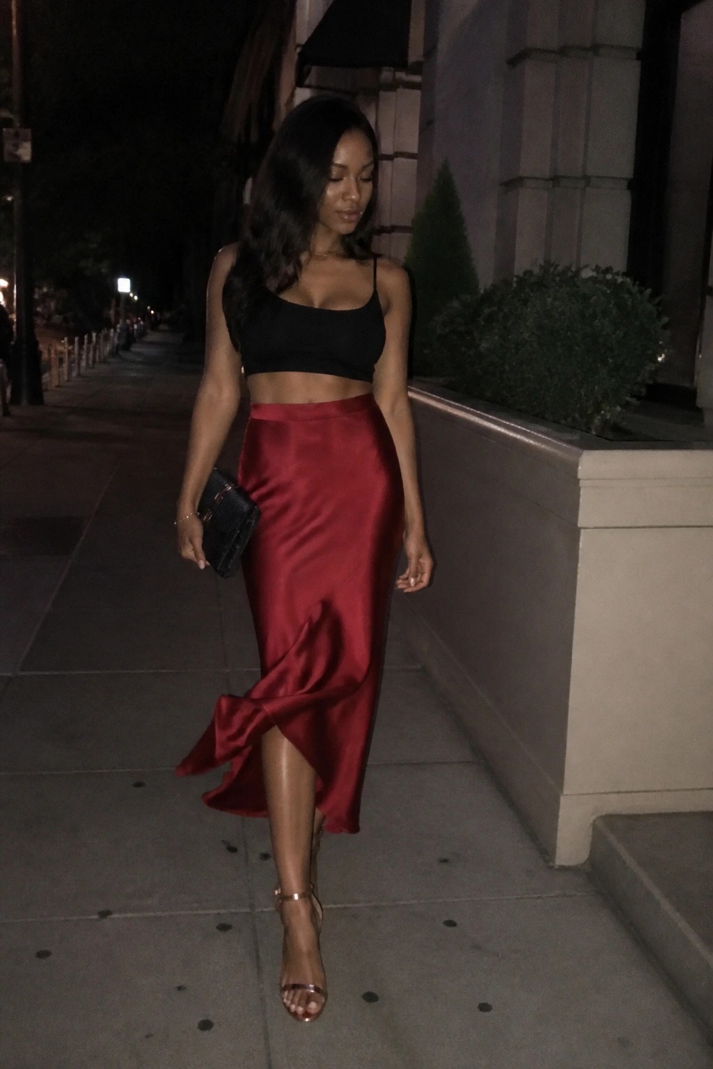 Woman in black crop top and flowing red satin skirt on nighttime city sidewalk