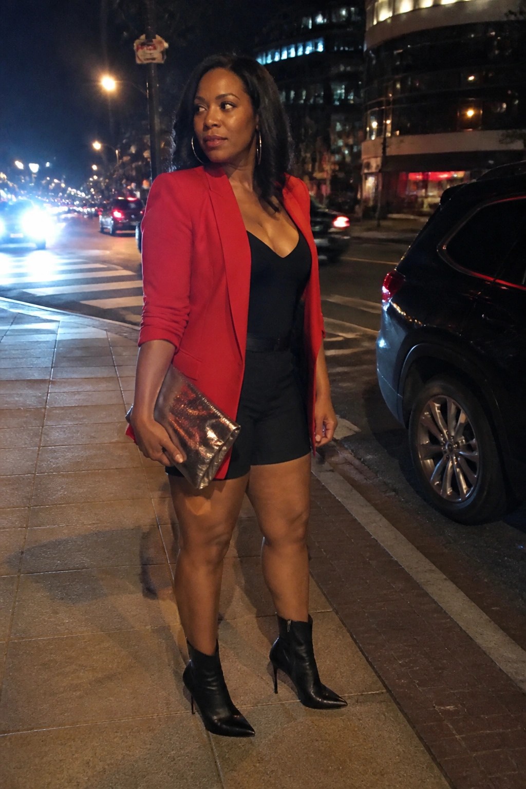 Woman in red blazer, black shorts, and heels holding metallic clutch on city street at night