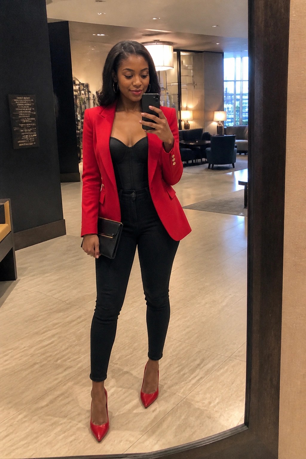 Woman wearing a red blazer over a black corset top with black jeans and red pointed heels while holding a black clutch in a hotel lobby mirror selfie.