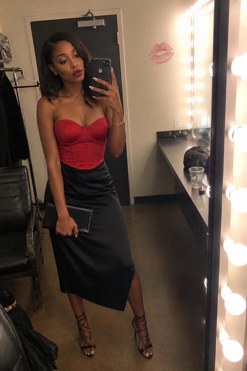 Woman in red corset top and black skirt takes mirror selfie in dressing room.