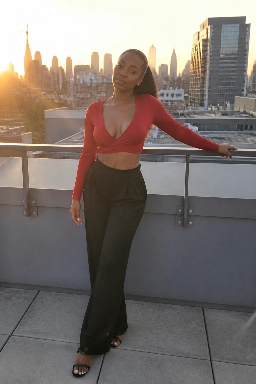 Woman in red crop top and black pants on balcony overlooking city skyline at sunset