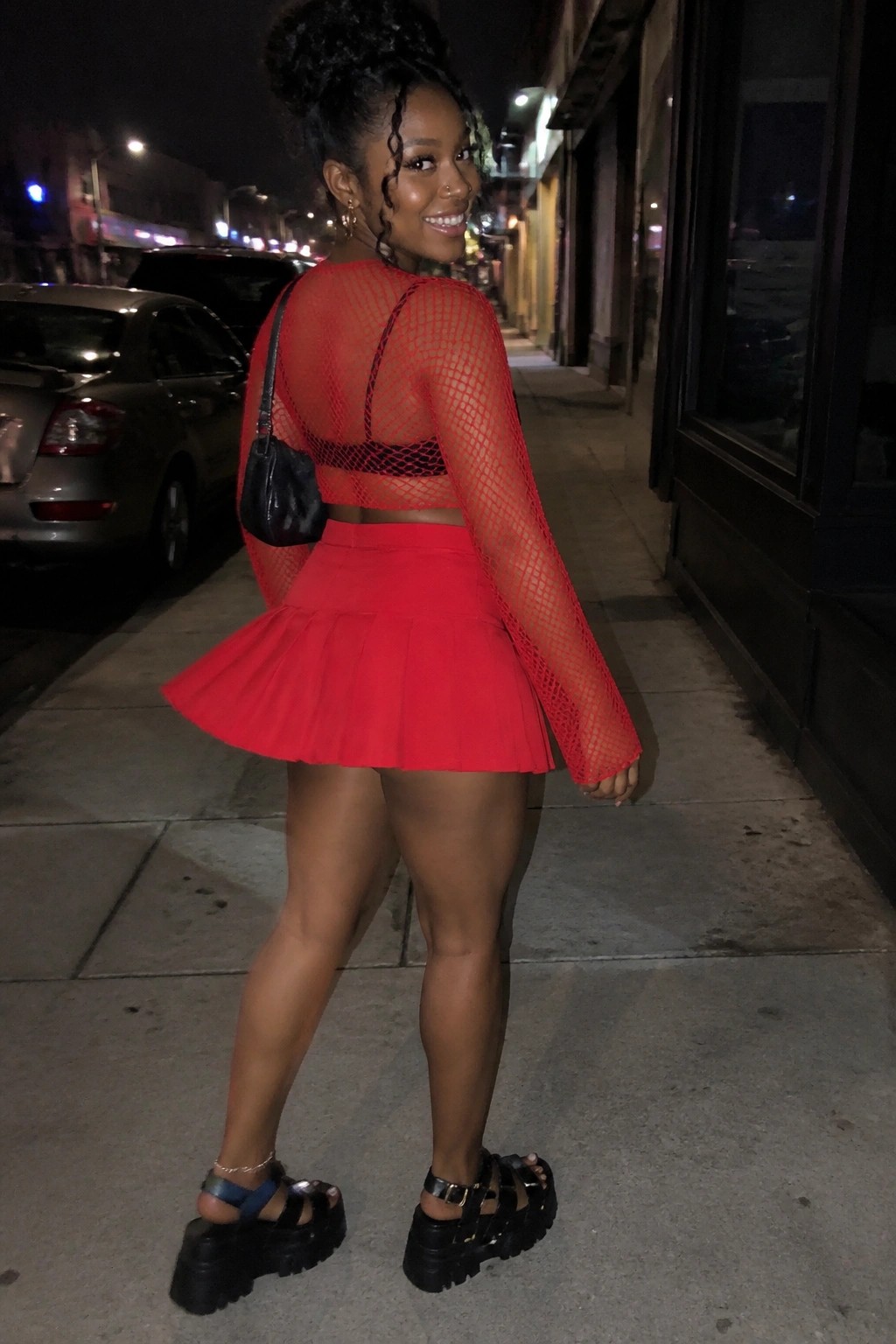 Woman in red fishnet crop top and pleated skirt on nighttime city sidewalk