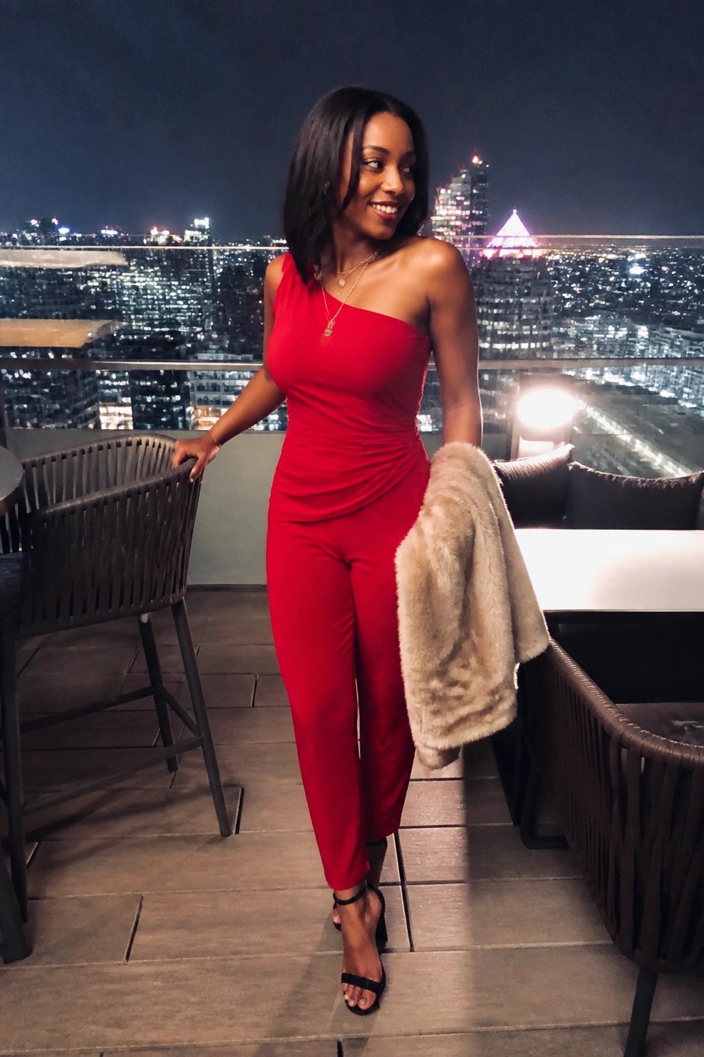 Woman in red one-shoulder jumpsuit holding beige faux fur coat with black heels and layered gold necklaces against night city view.