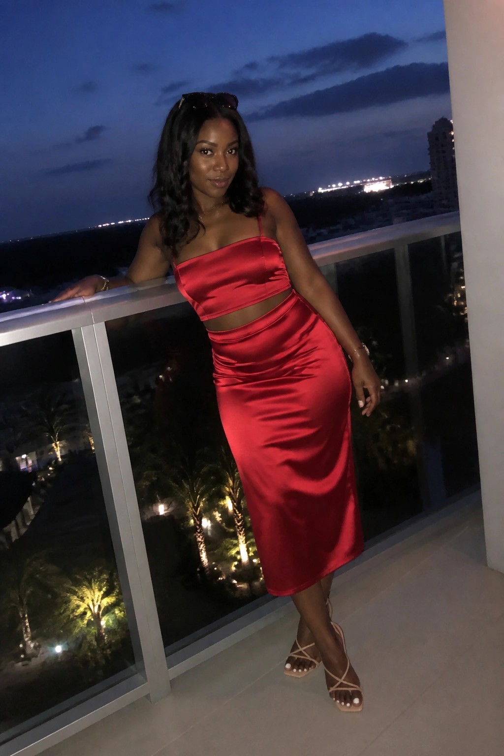 Woman wearing a red satin strapless midi dress with waist cutout and strappy beige heels standing on a balcony at night.