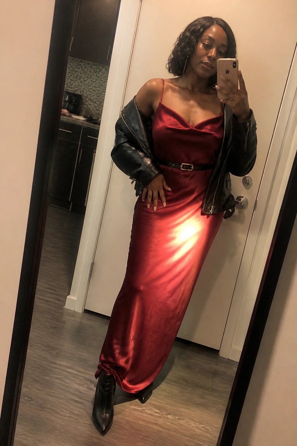 Woman in shiny red satin dress with black leather jacket taking mirror selfie.
