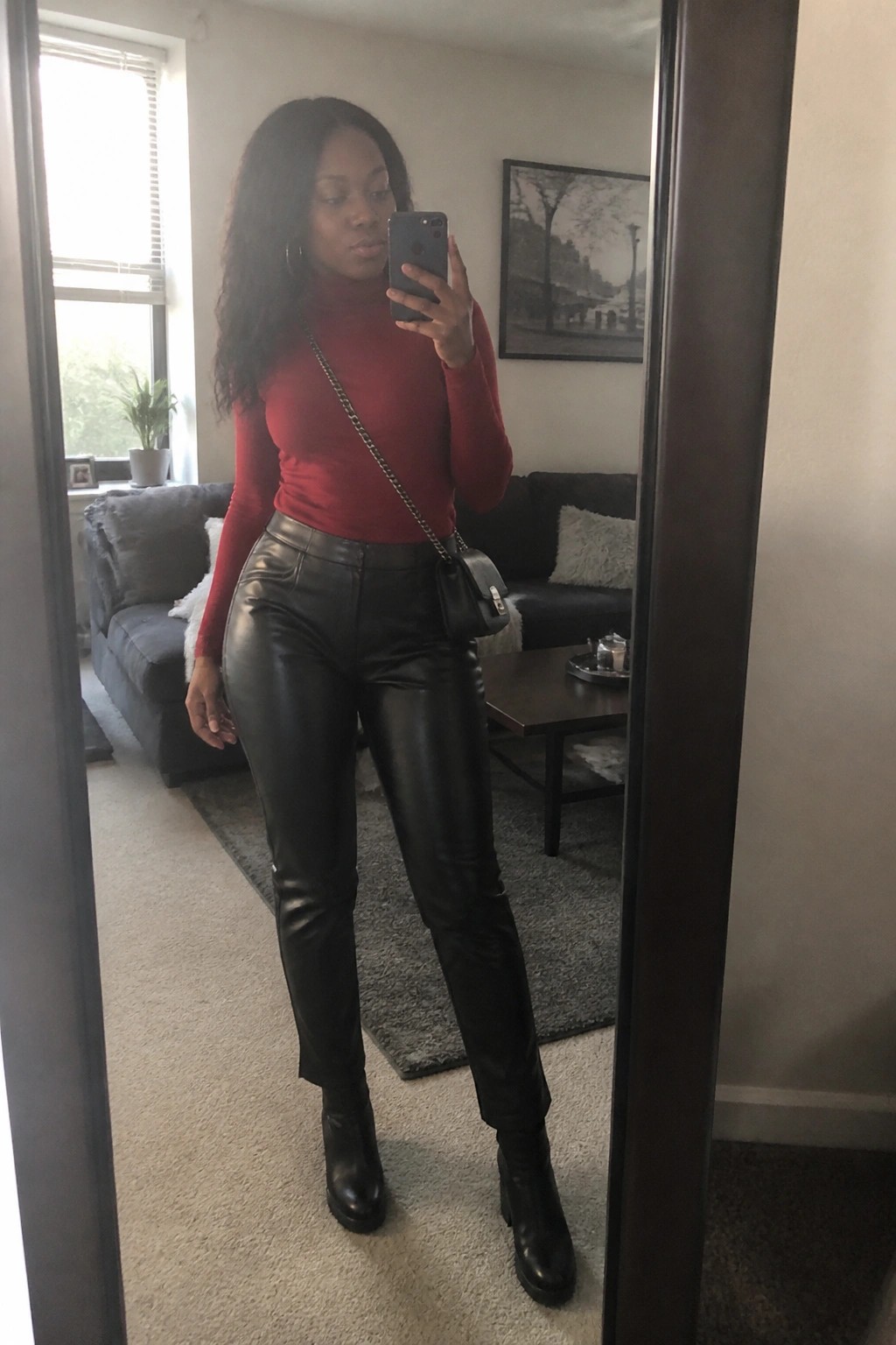 Woman in red turtleneck and black leather pants taking mirror selfie in living room