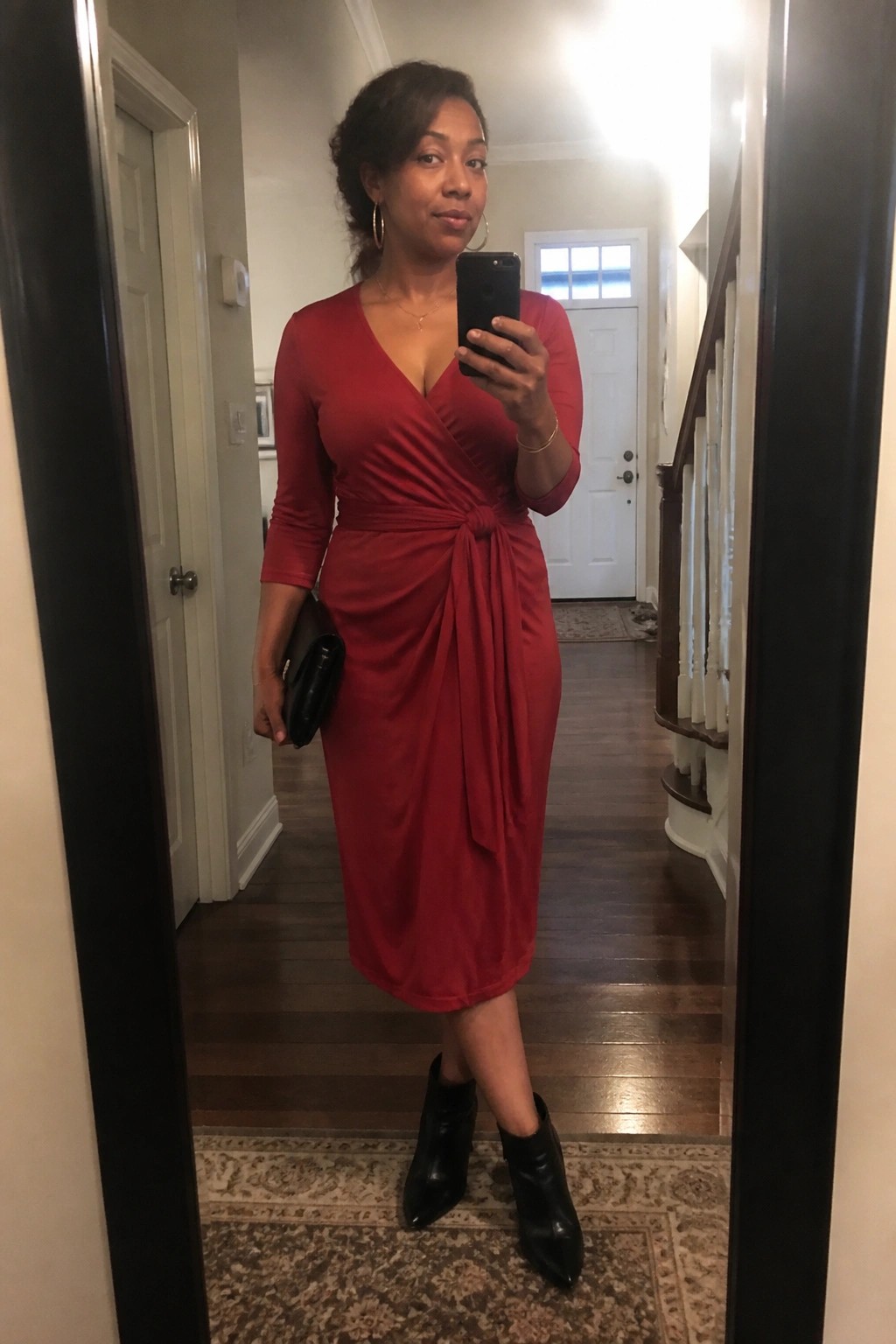 Woman in red wrap midi dress with knot tie and black ankle boots taking a mirror selfie while holding a clutch.