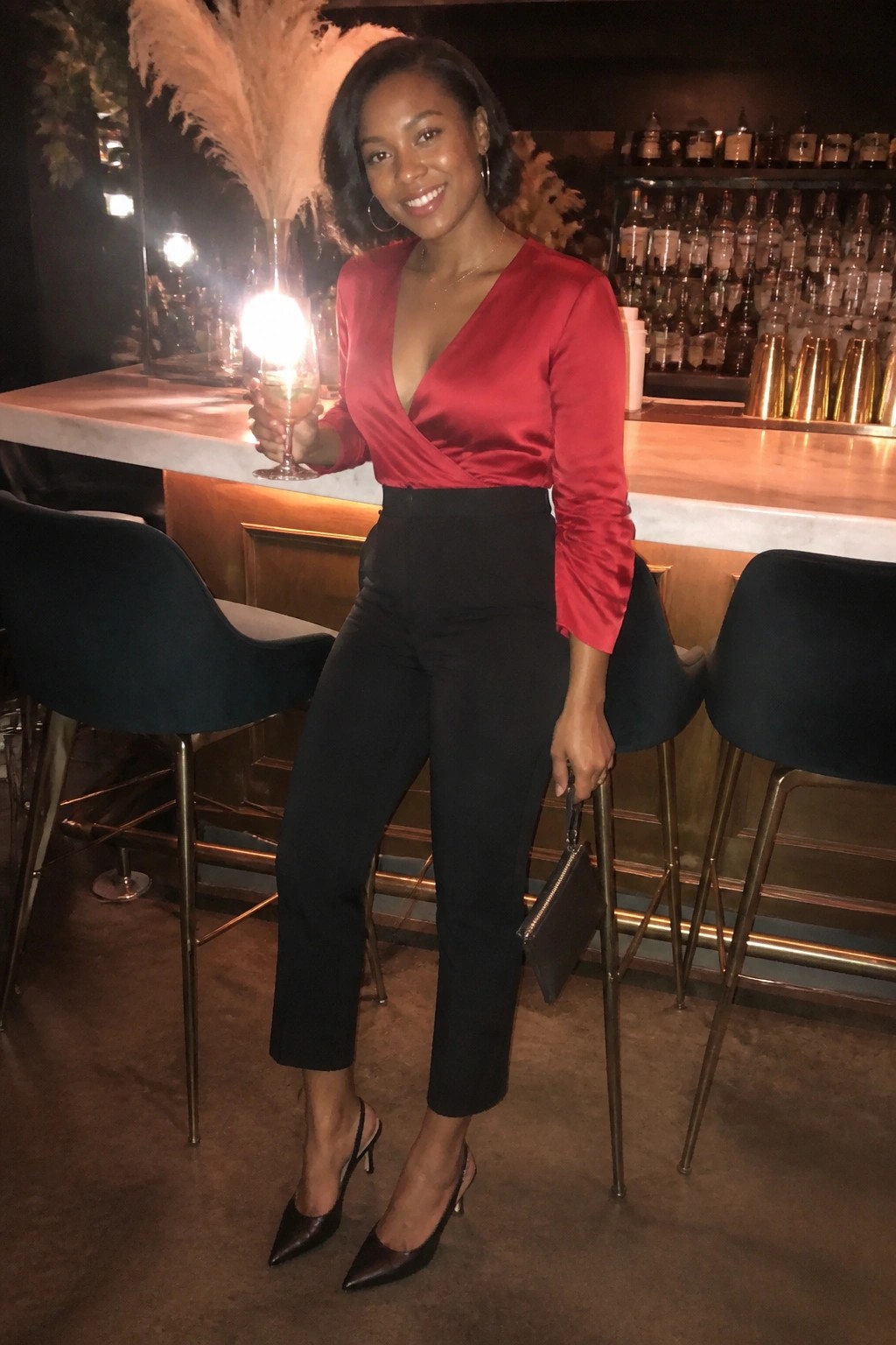 Woman in red satin wrap blouse tucked into black cropped trousers with pointed black heels and a small clutch.