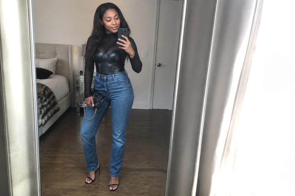19 Luxe Leather Corset Outfit Ideas for Black Women That Elevate Night Out Style Woman in black mesh top and leather corset with blue jeans taking mirror selfie in bedroom