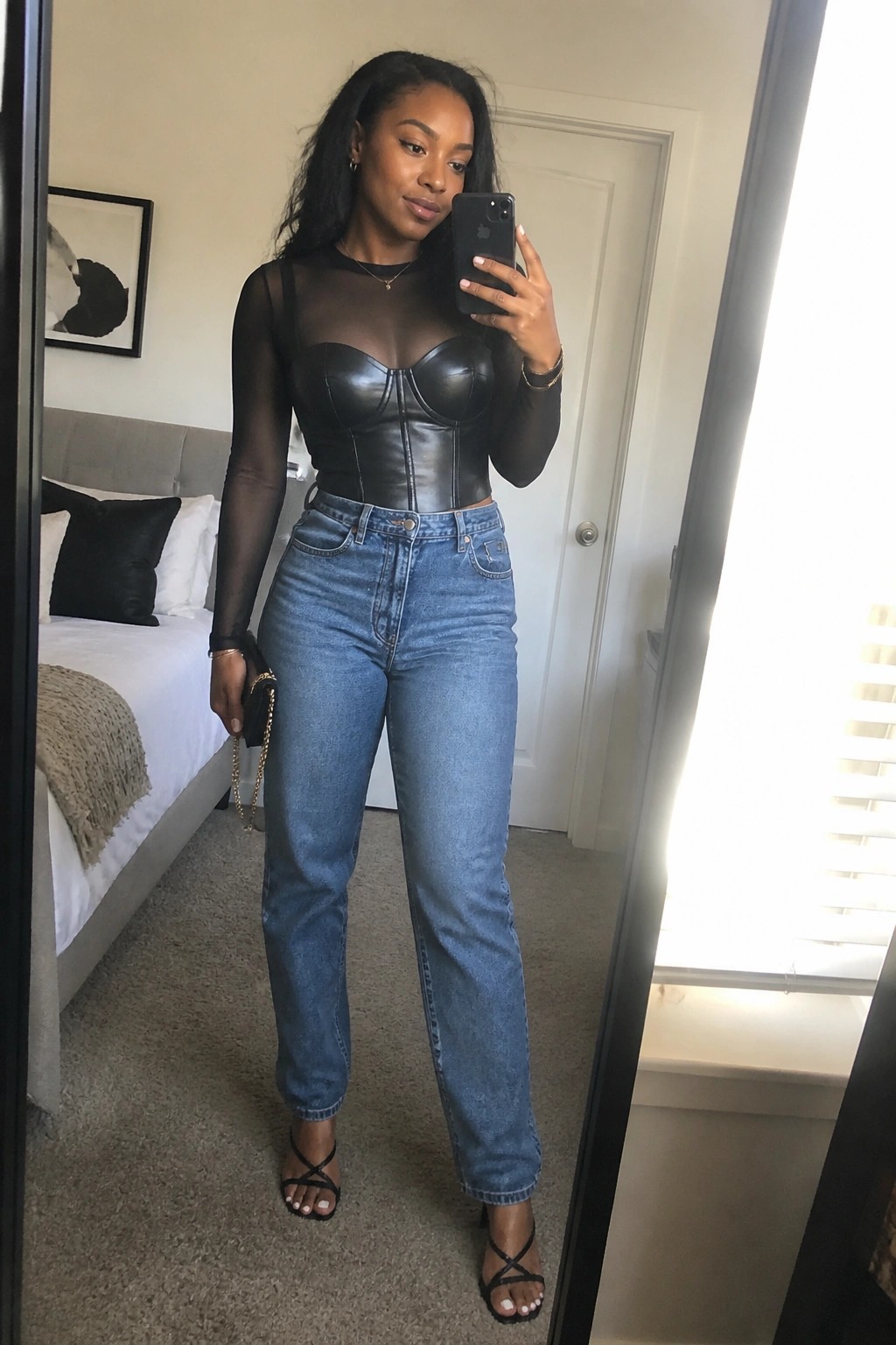 Woman in black mesh top and leather corset with blue jeans taking mirror selfie in bedroom