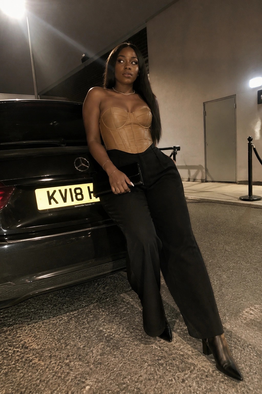 Woman in tan corset top and black pants leaning on black Mercedes at night
