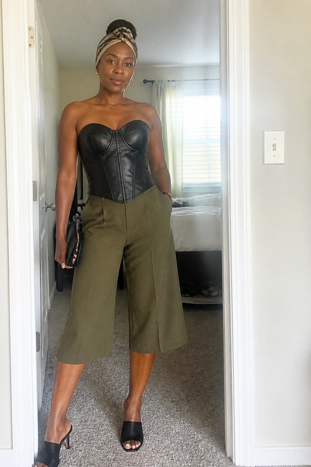 Woman in black leather corset, olive culottes, and patterned headwrap in doorway.