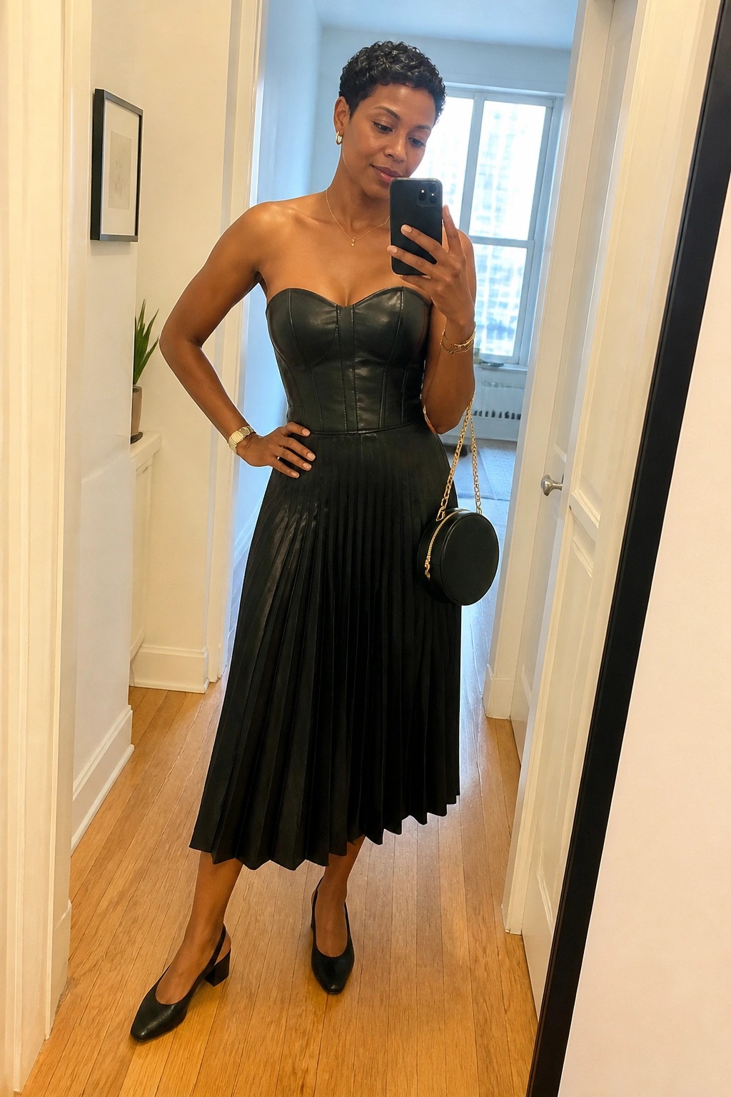 Woman in black strapless pleated leather dress taking mirror selfie with phone and purse.