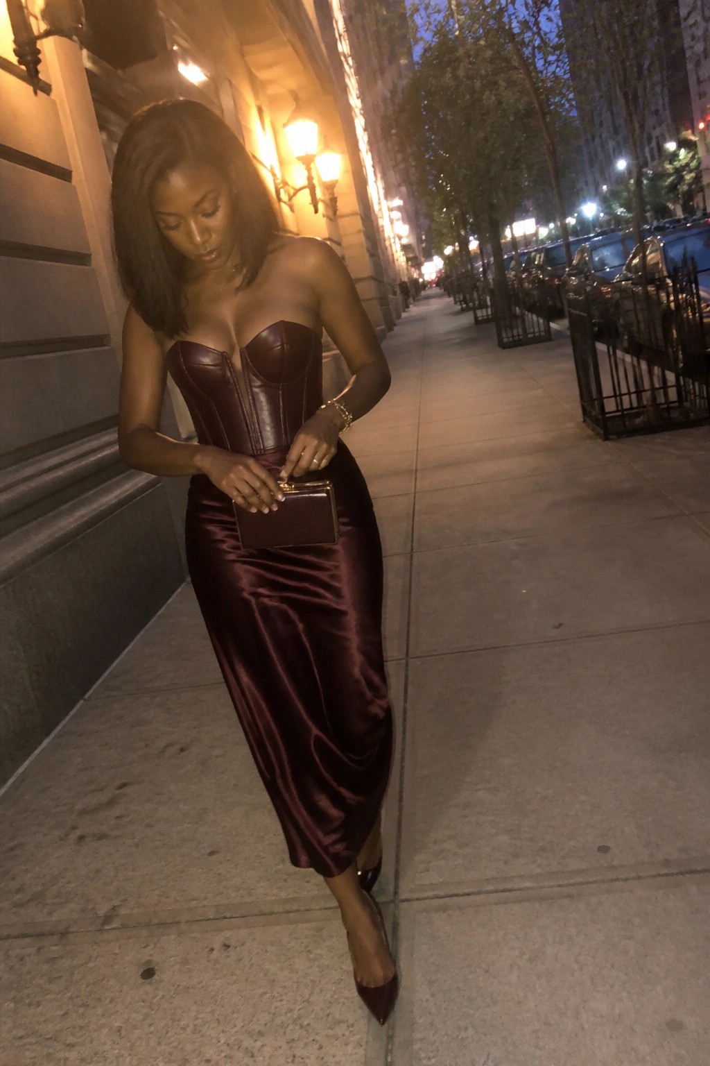 Woman in maroon strapless corset top and satin skirt holding clutch on city sidewalk at night