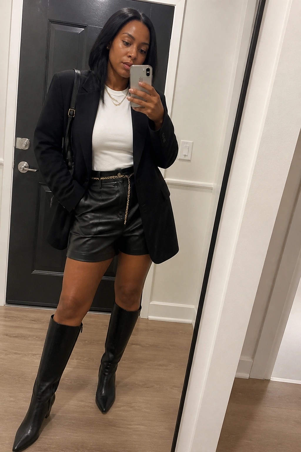 Woman in black blazer, white top, and leather shorts holds phone in mirror selfie.