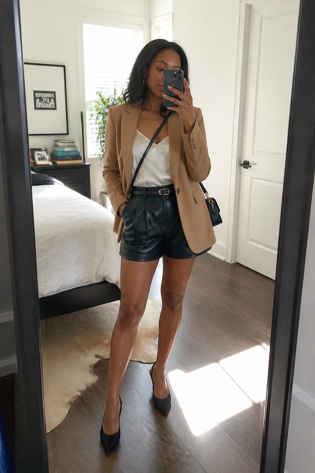 Woman in tan blazer and black leather shorts taking mirror selfie in bedroom