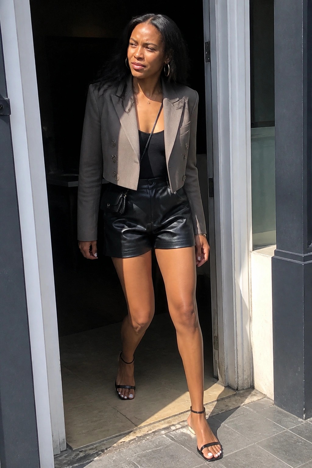 Woman in cropped gray blazer and black leather shorts stands in sunlit doorway.