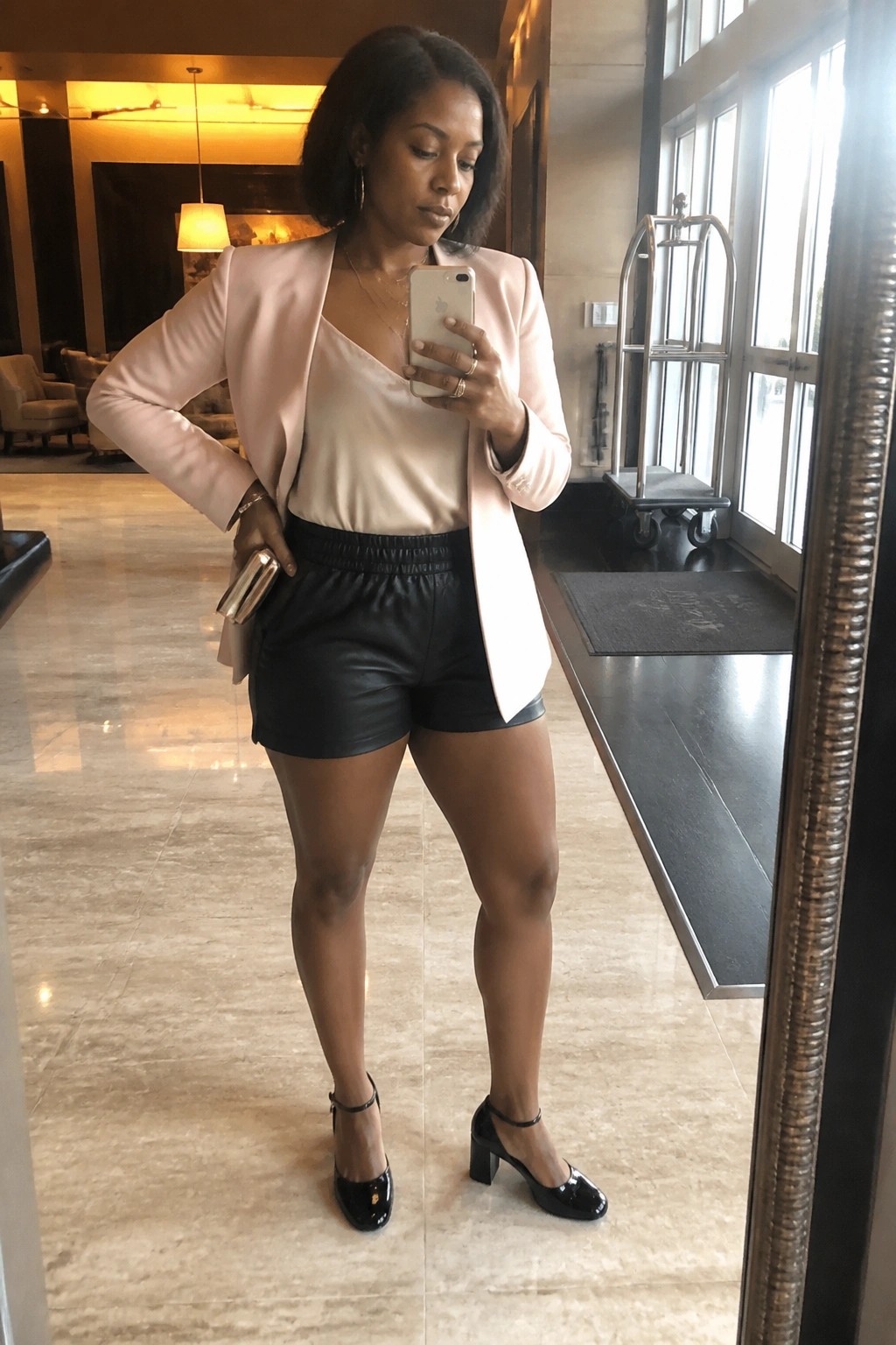 Woman in light pink blazer and black leather shorts taking mirror selfie in elegant lobby.