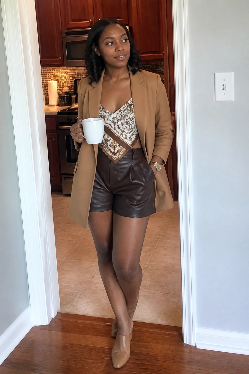Woman in tan blazer, patterned top, and brown shorts holding white mug in doorway
