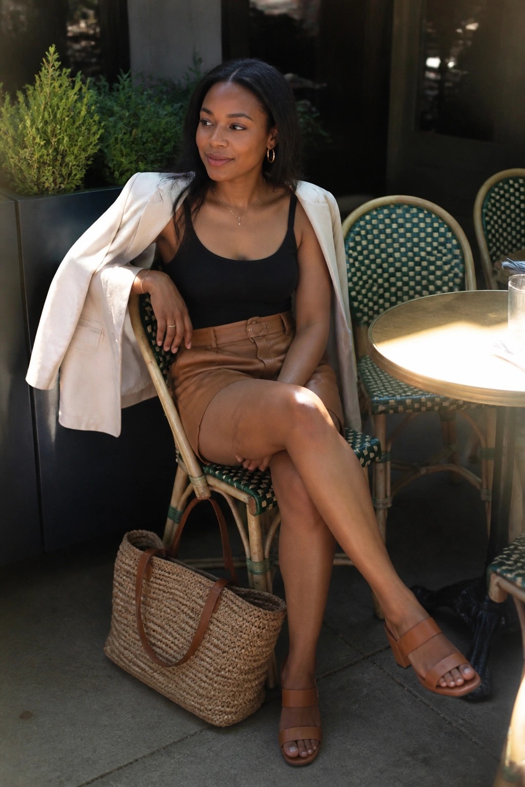 Woman in black top, brown leather skirt, and beige blazer at outdoor cafe with straw bag