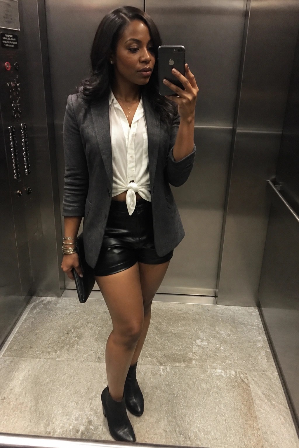 Woman in gray blazer, tied white blouse, and black leather shorts takes elevator selfie.