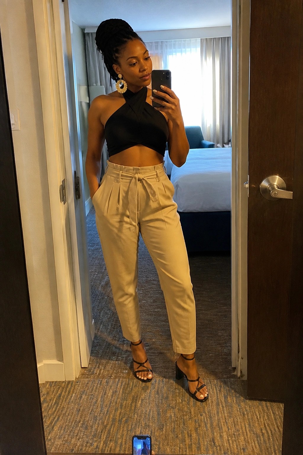 Woman in black halter crop top and beige pants taking mirror selfie in hotel room