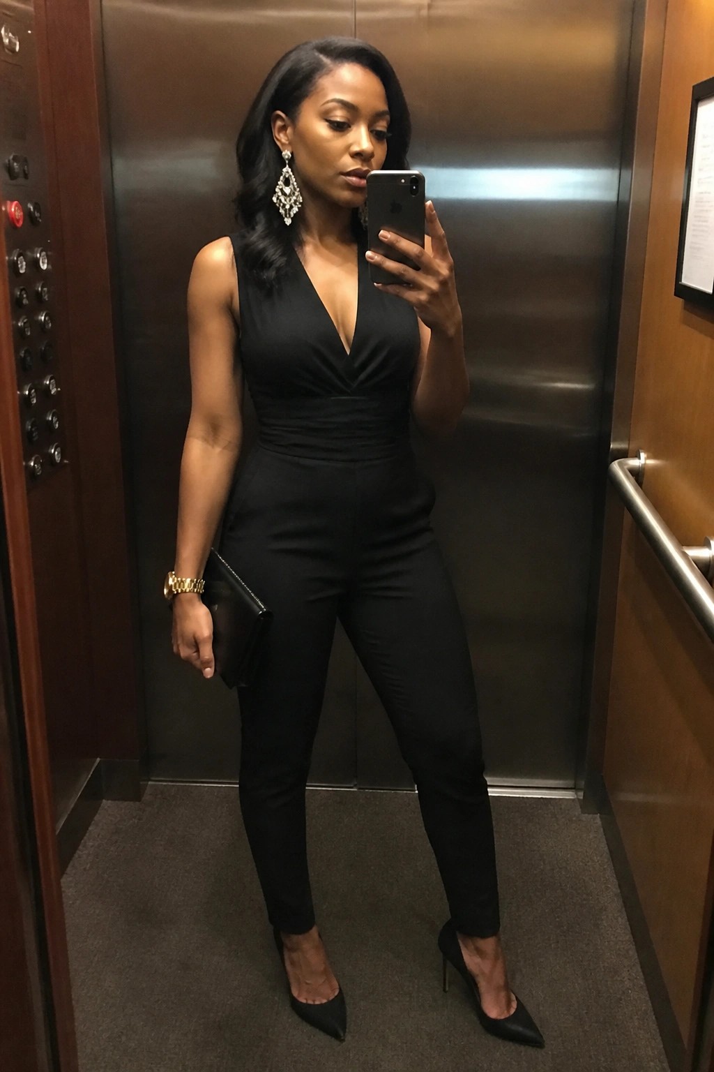 Woman wearing a fitted sleeveless black jumpsuit with deep V neckline, black pointed toe heels, holding a black clutch and wearing large dangling earrings while taking a mirror selfie.