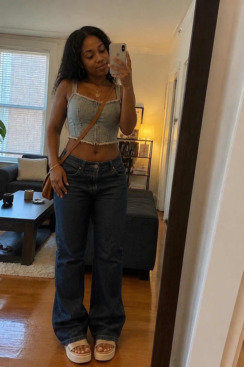 Woman in light blue denim crop top with zipper front and dark wide leg jeans paired with white platform slides and brown crossbody bag standing in a living room taking a mirror selfie.