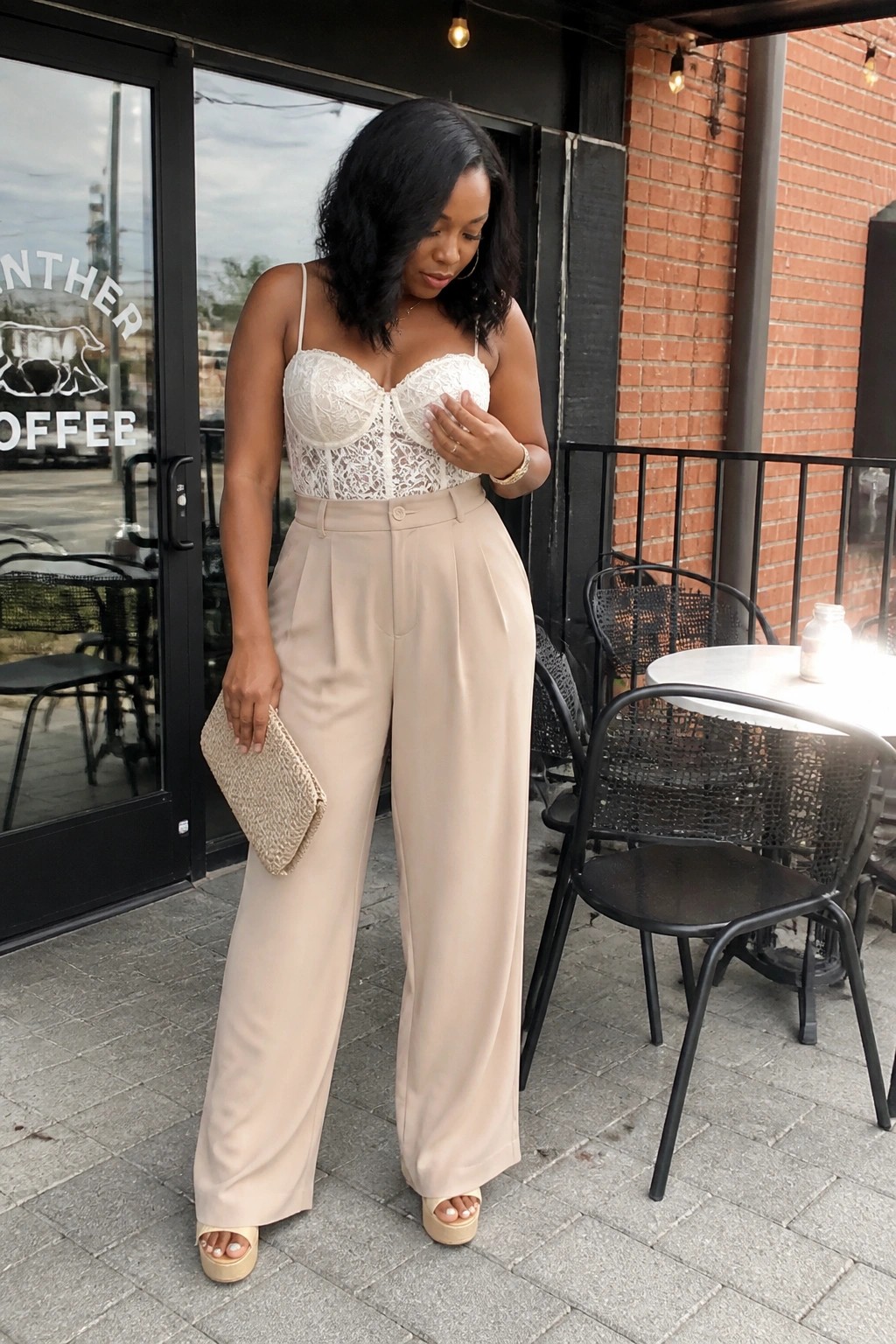 Woman in white lace corset top and beige wide-leg pants holding clutch at cafe
