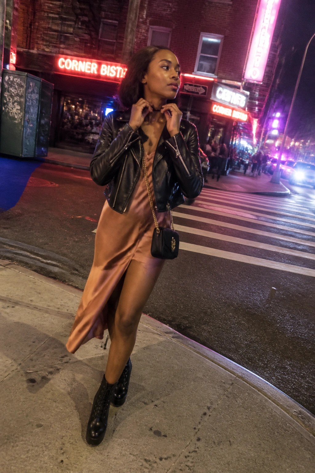 Woman in black leather jacket and satin dress adjusts necklace on neon city street