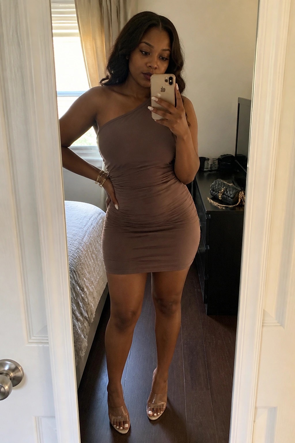 Woman wearing a taupe one-shoulder ruched mini dress with clear heeled sandals while taking a mirror selfie in a bedroom.