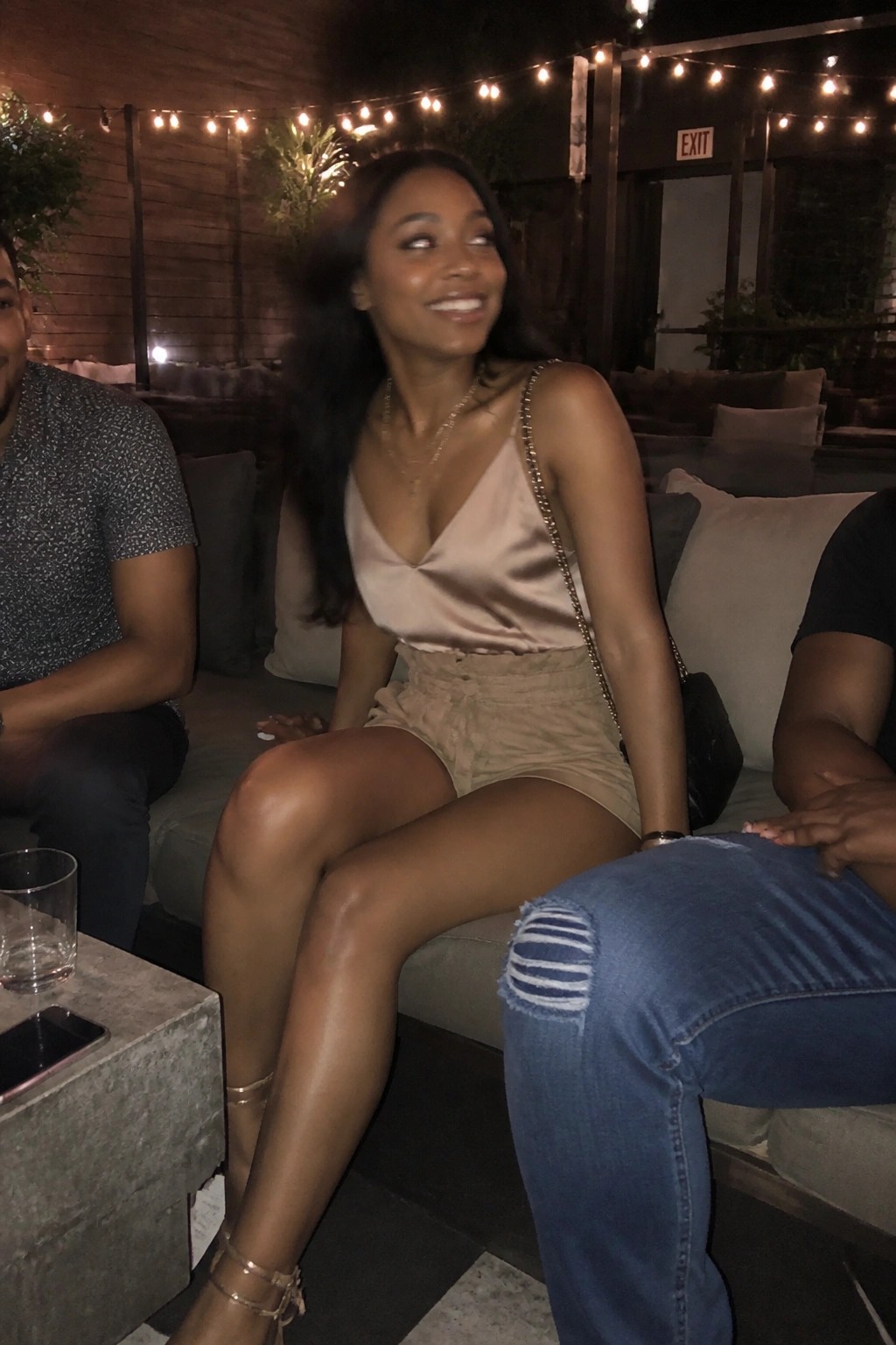 Woman in beige satin top and shorts smiling on couch under string lights at night