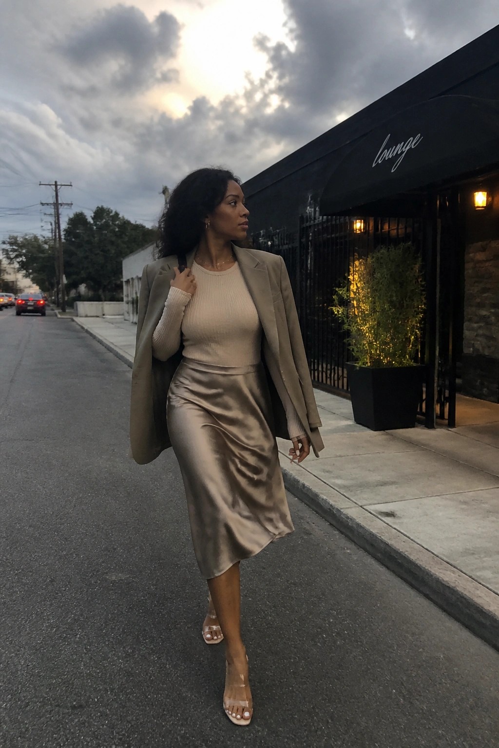Woman in beige blazer and satin skirt walks past black lounge awning at dusk