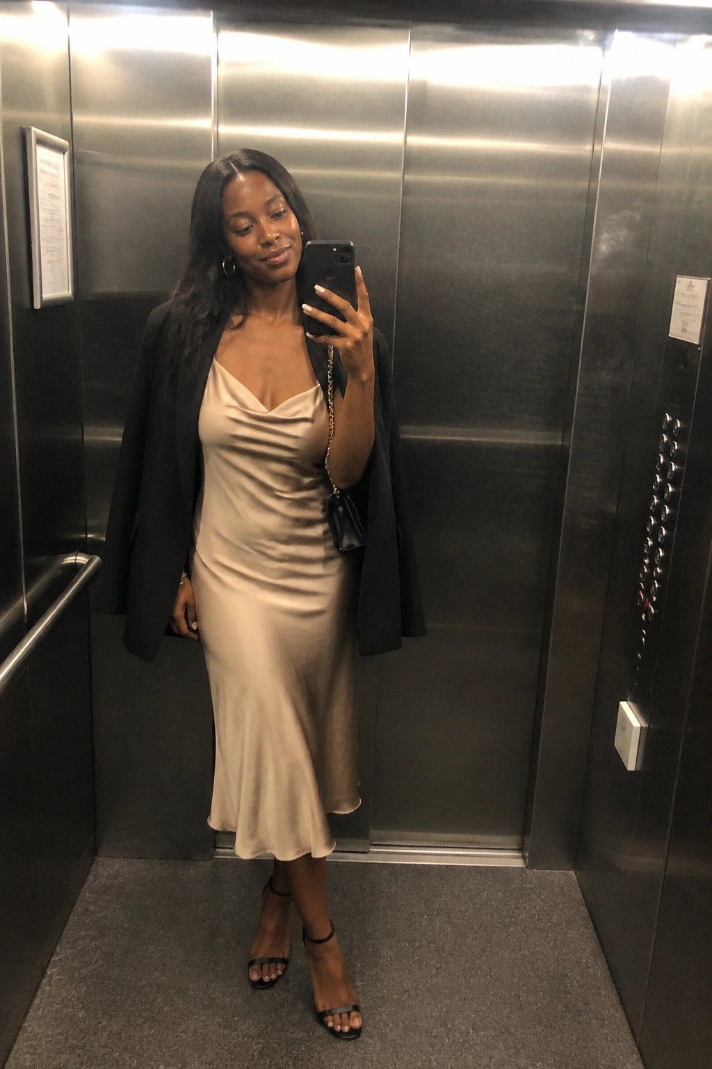Woman in beige satin dress and black blazer taking selfie in elevator