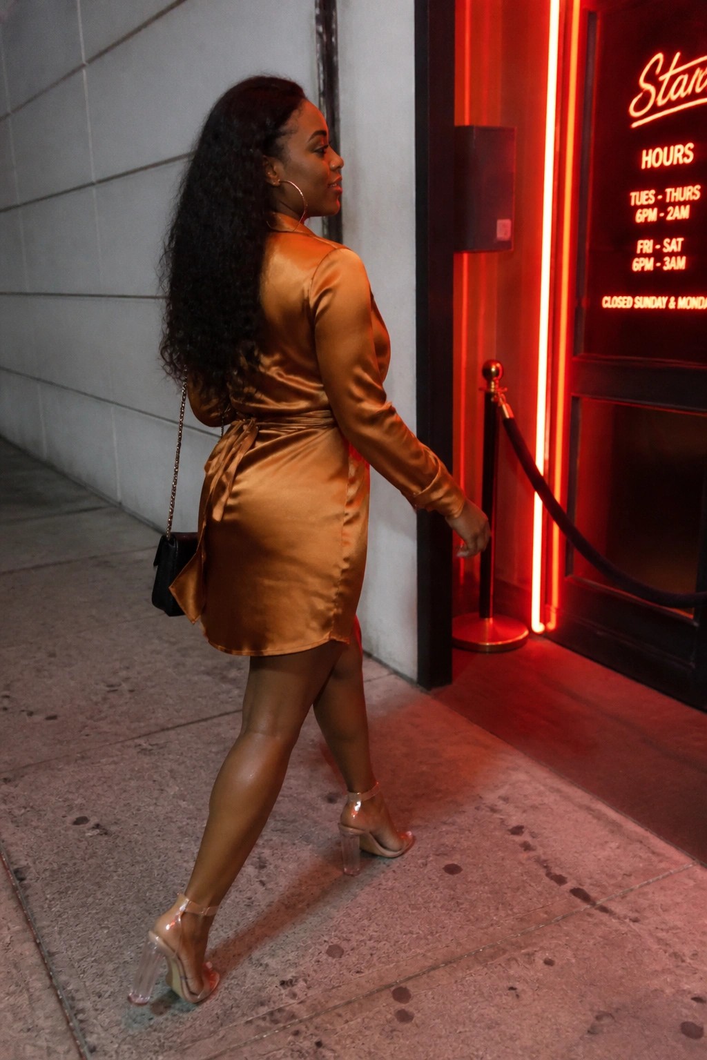 Woman in orange satin dress and clear heels walks past red neon nightclub entrance.