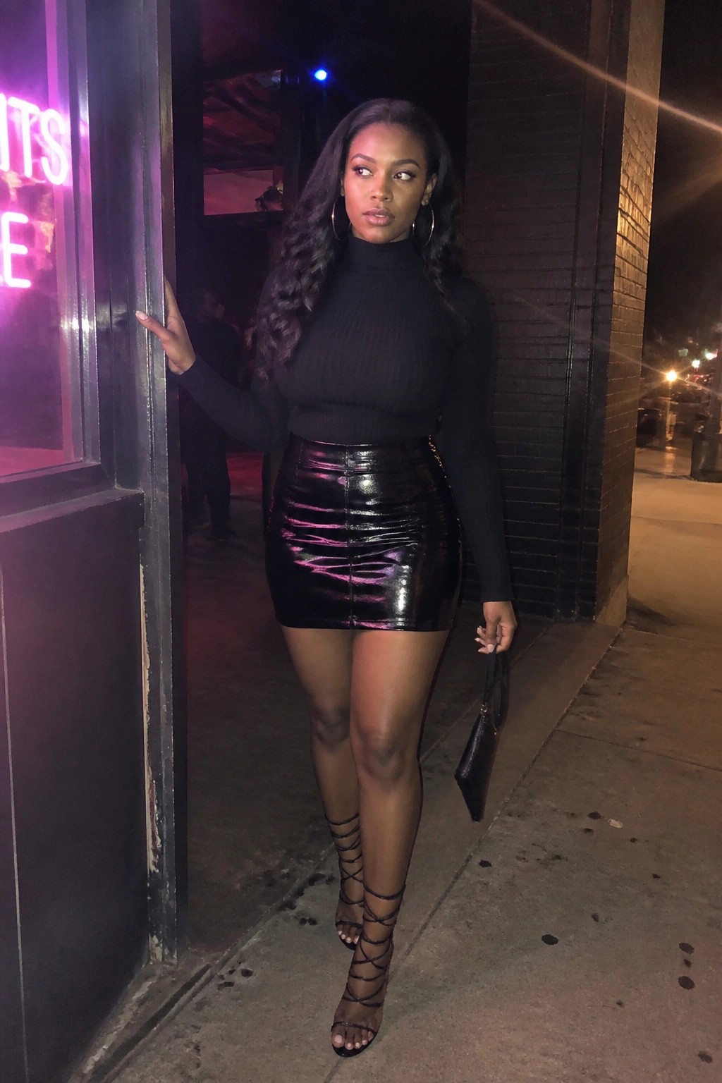 Woman wearing a fitted black ribbed turtleneck sweater tucked into a glossy black mini skirt with strappy black high heels and large hoop earrings while holding a small black purse.