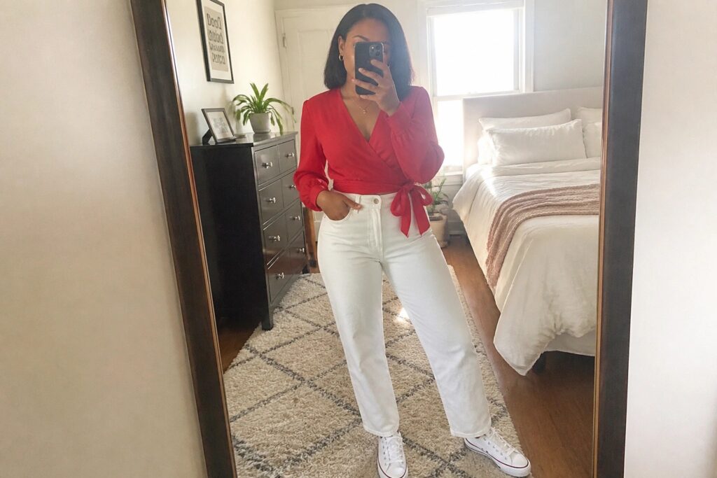 Woman in red wrap blouse and white jeans taking mirror selfie in bedroom