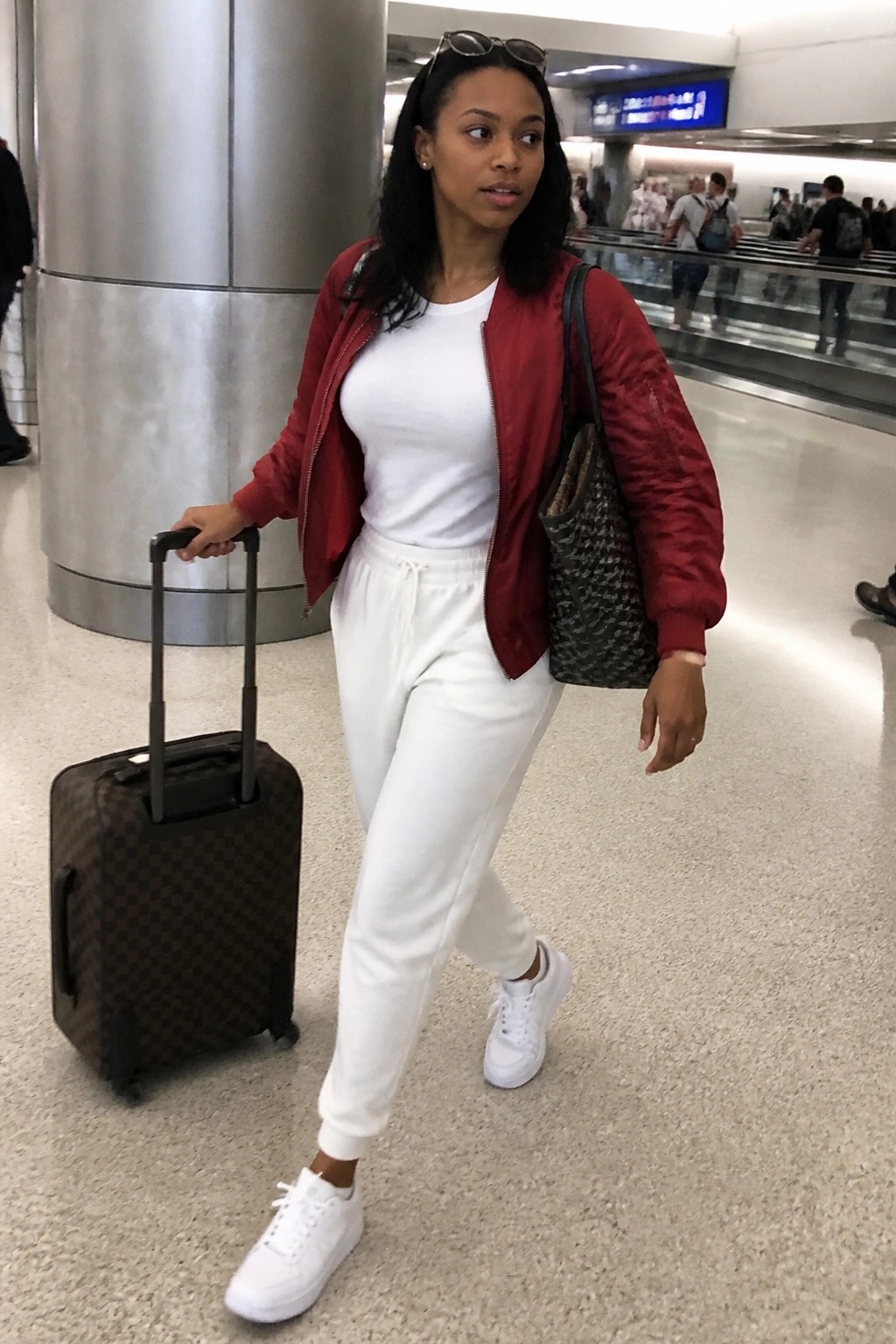 Woman wearing an open red bomber jacket over a white t-shirt and white joggers with white sneakers while pulling a rolling suitcase in an airport terminal.
