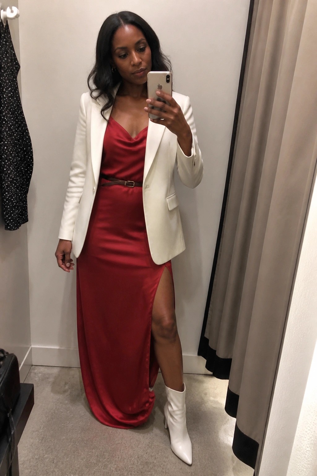 Woman in red satin dress and white blazer taking mirror selfie in fitting room