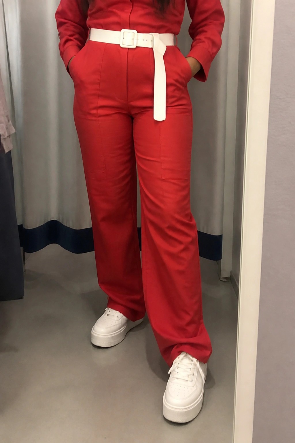 Person in red shirt and pants with white belt and platform sneakers, hands in pockets.