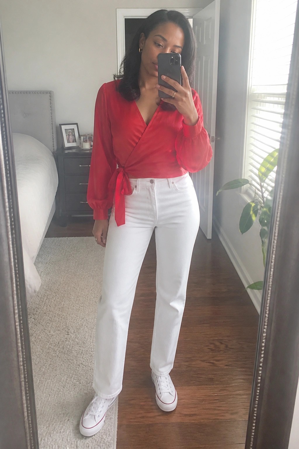 Woman in red wrap blouse and white jeans taking mirror selfie in bedroom