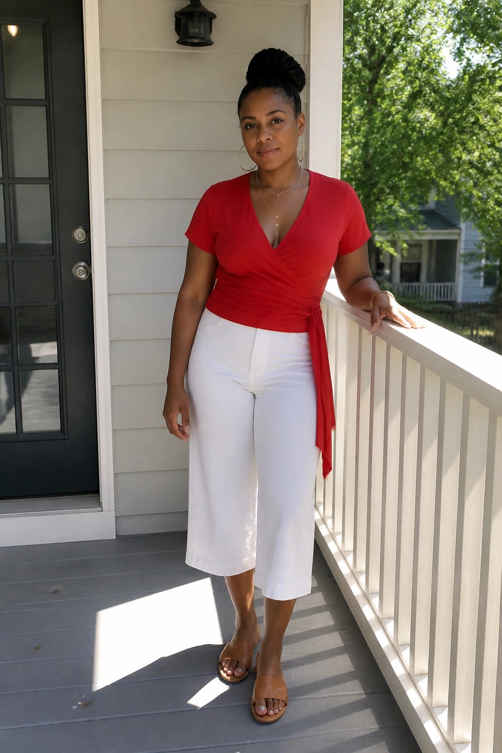 Woman in red wrap top and white capris leaning on sunny porch railing.