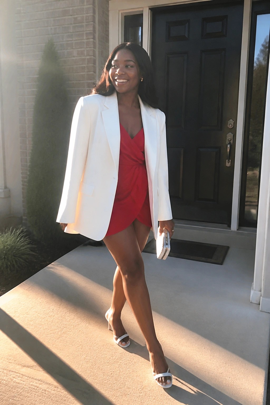 Woman in white blazer over red mini dress on sunny house porch holding clutch