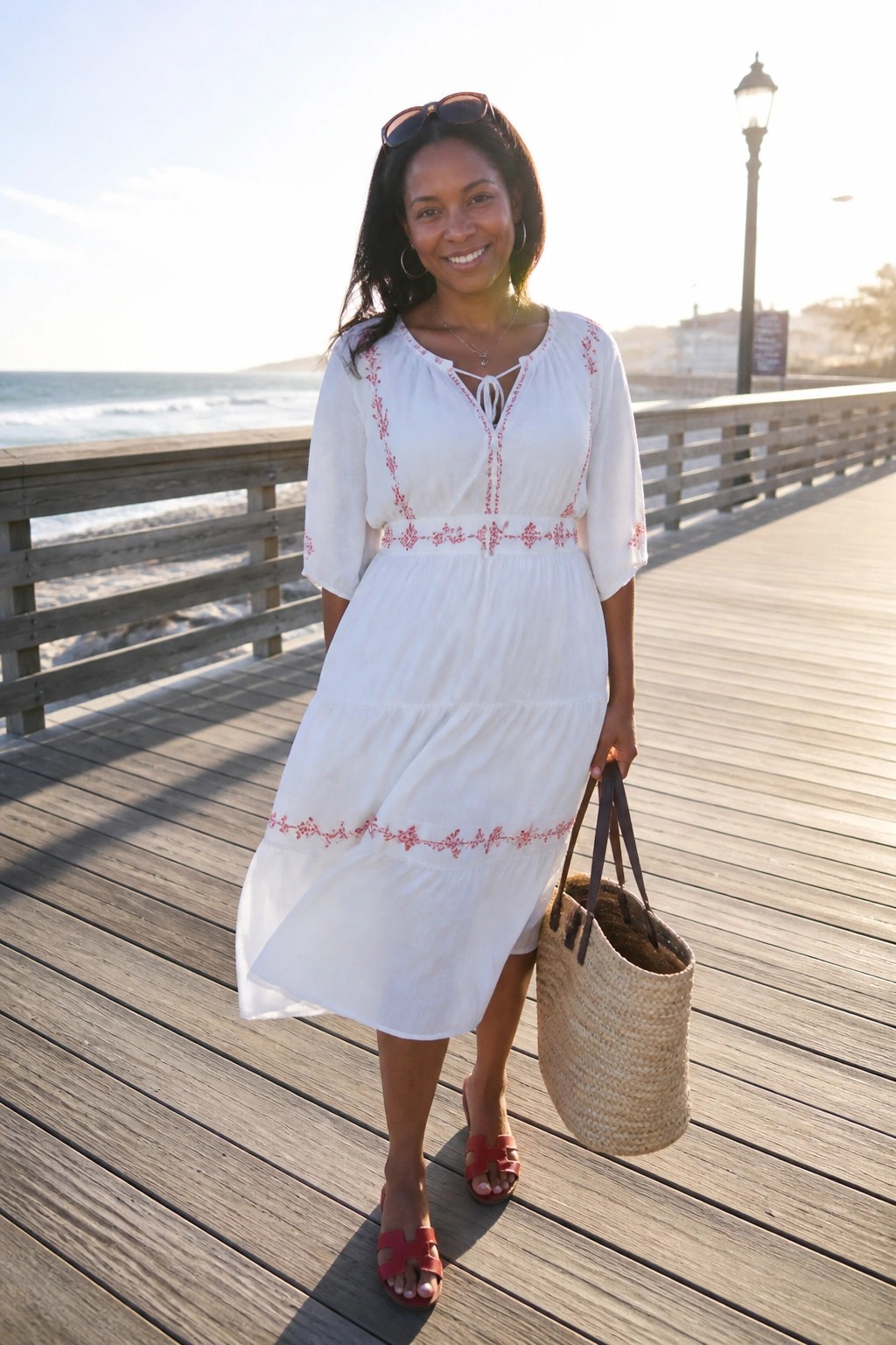 Woman in white tiered dress with red embroidery holding straw bag on ocean pier