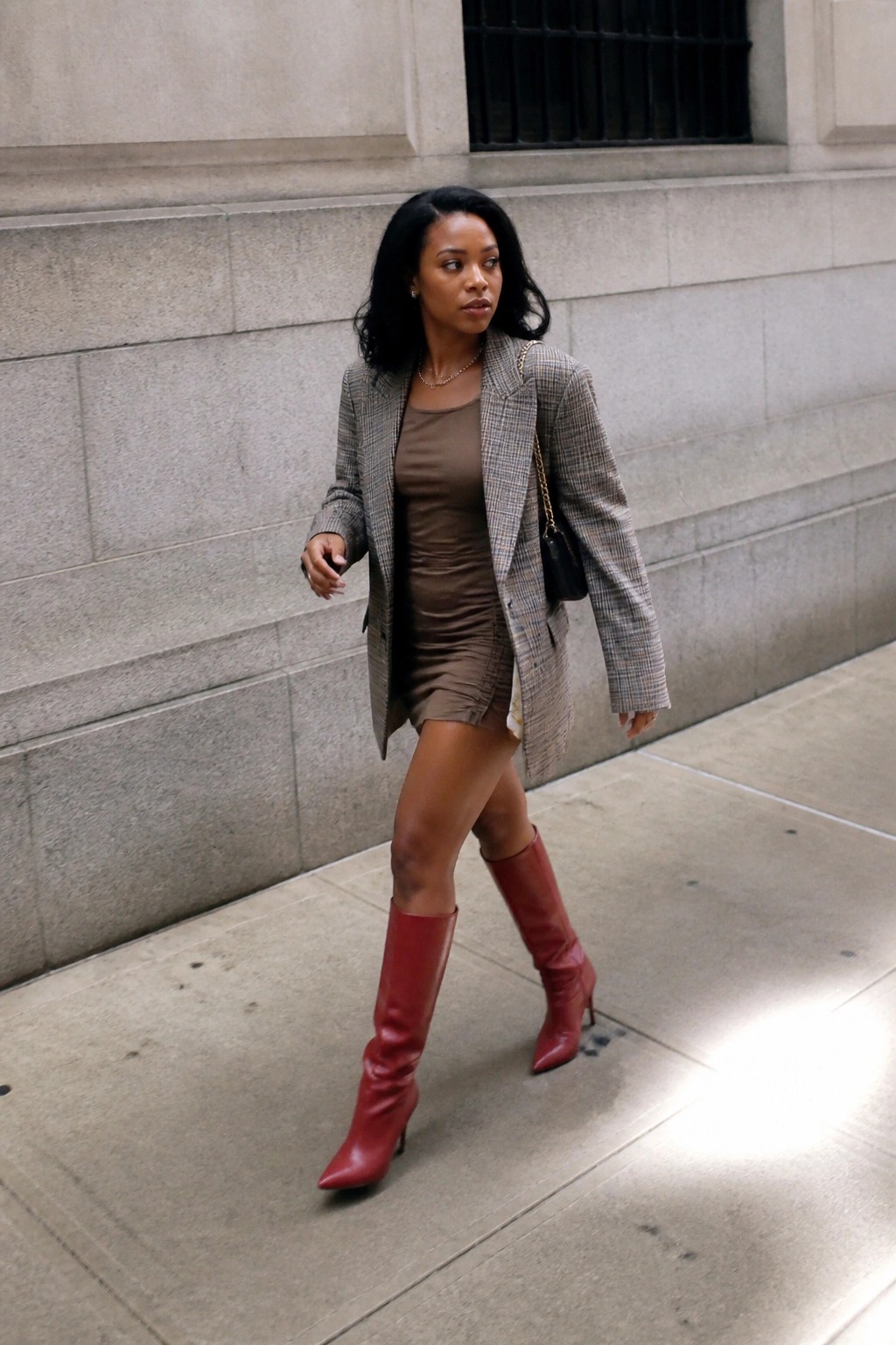 Woman in plaid blazer, brown dress, and red knee-high boots walking on sidewalk