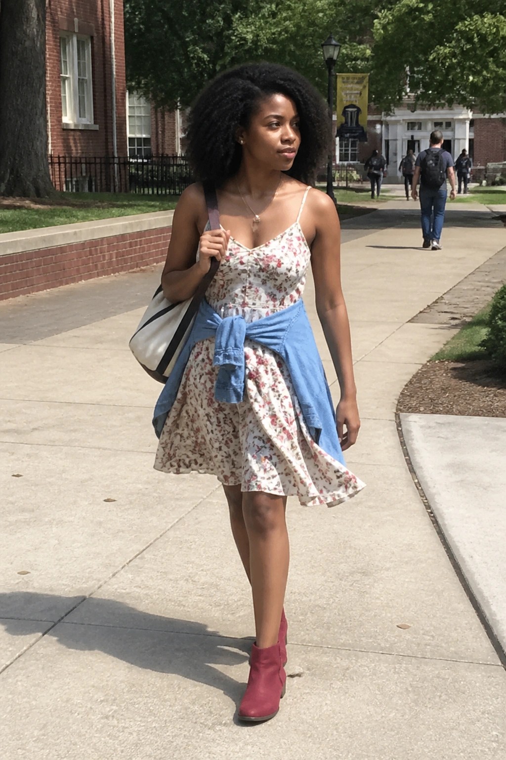 Black woman wearing a white floral print sundress with thin straps, a blue denim jacket tied around the waist, red ankle boots, and carrying a large tote bag on a paved walkway.