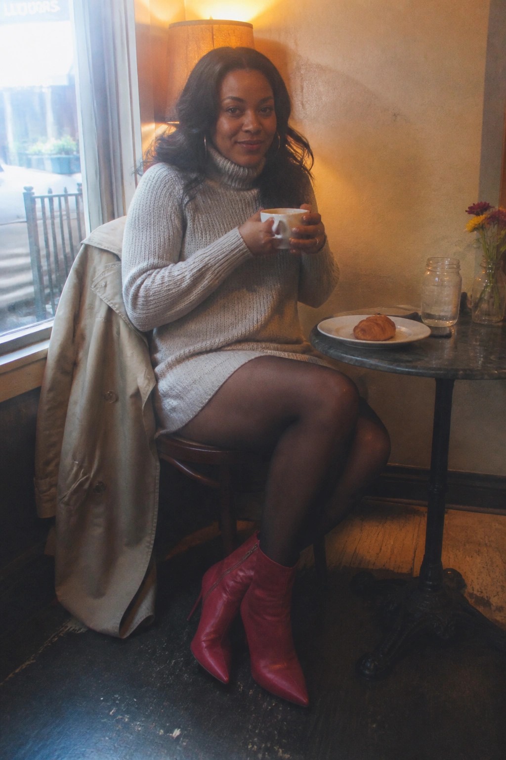 Woman in gray turtleneck dress and red boots holding cup beside croissant