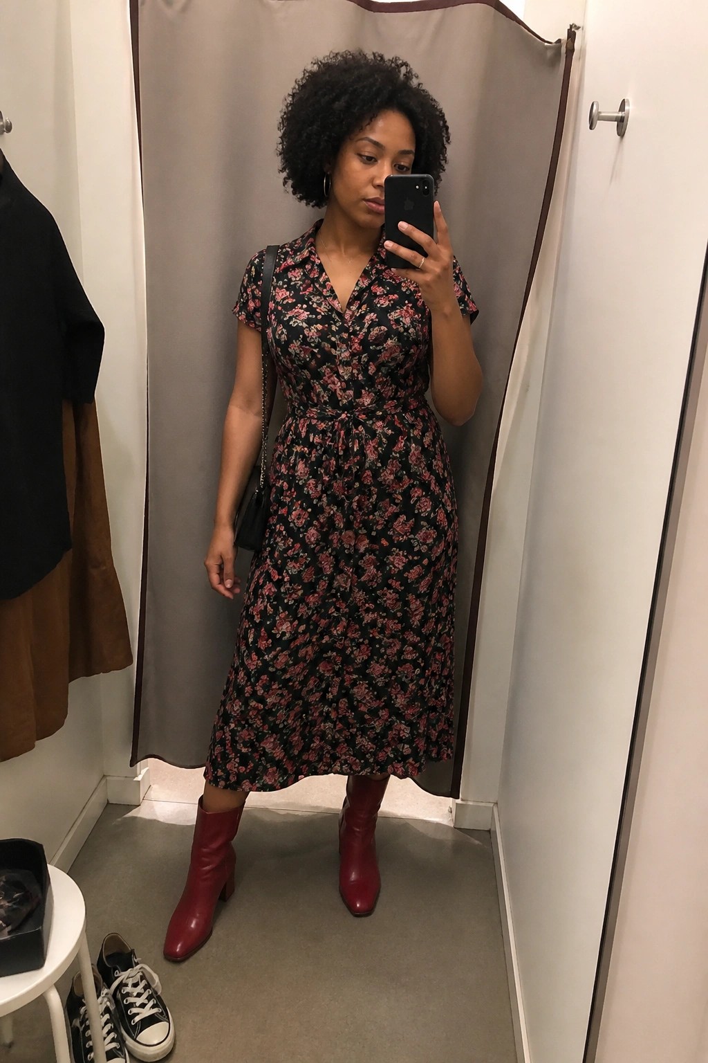 Curly-haired woman in black floral midi dress and red boots taking mirror selfie.