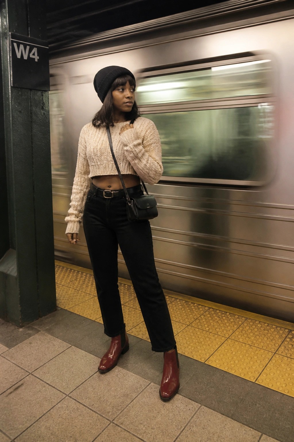Woman in cream cropped sweater, black beanie, and red boots on subway platform with passing train